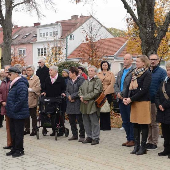 Eine Gruppe von Menschen, einige davon ältere Personen, steht in einem Außenbereich vor einem Gebäude. Sie stehen in einer Reihe und warten möglicherweise auf etwas.