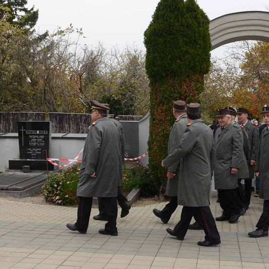 Eine Gruppe uniformierter Personen geht auf einem Friedhof mit einem Gedenkstein im Hintergrund.