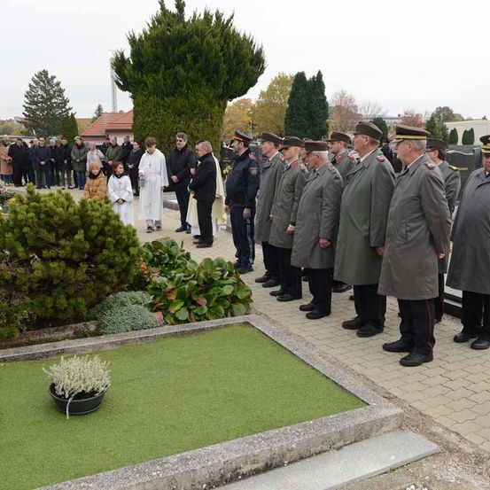 Eine feierliche Zeremonie auf einem Friedhof, mit uniformierten Personen in Reihen, einem Priester in einer weißen Robe und einer Gruppe von Zuschauern. Grüne Pflanzen und ein kleiner Blumentopf im Vordergrund.