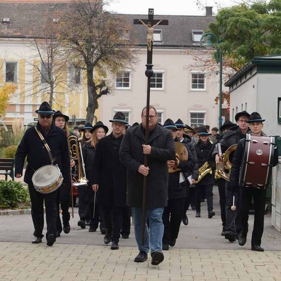 Eine Gruppe von Menschen, die marschieren, Musikinstrumente spielen und ein Kreuz auf einem Stab tragen, mit Gebäuden und Bäumen im Hintergrund.