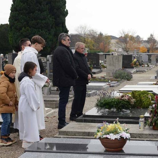 Eine Gruppe von Menschen, einschließlich Kinder, steht auf einem Friedhof, in der Nähe eines Grabes. Sie tragen Winterkleidung. Es gibt Blumen und Kerzen auf dem Grab.