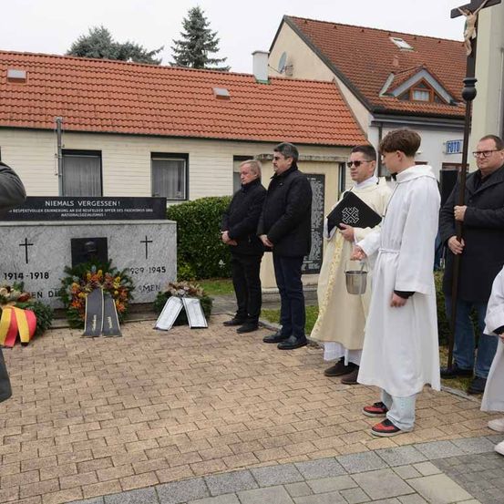Eine Gruppe von Männern in formeller Kleidung und ein Priester in einer weißen Robe stehen vor einem Denkmal, halten einen Eimer und ein Buch. Das Denkmal hat ein Kreuz und den Text 'NIEMALS VERGESSEN!'.