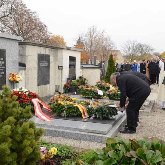 Ein Mann in einem schwarzen Anzug legt Blumen an einem Grab an, wo sich viele Grabsteine und Blumen befinden.