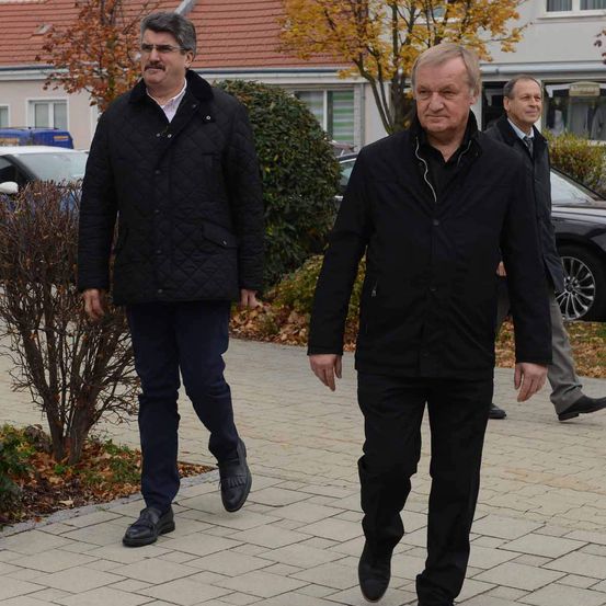 Drei Männer gehen auf einem gepflasterten Bürgersteig. Ein Mann führt, trägt einen schwarzen Mantel. Hinter ihm folgt ein weiterer Mann in einem ähnlichen Mantel. Ein dritter Mann in einem Anzug geht hinter ihnen. Sie stehen vor einem Gebäude mit einem roten Dach und geparkten Autos.