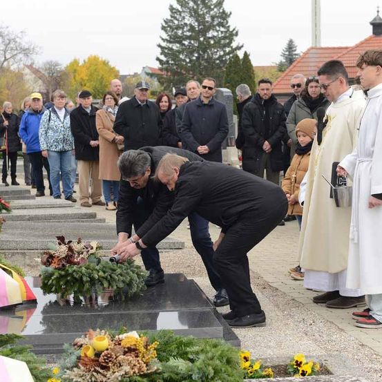 Eine Gedenkfeier, bei der mehrere Männer Kränze an einem Grabstein ablegen, mit Zuschauern und Priestern, bei klarem Himmel.