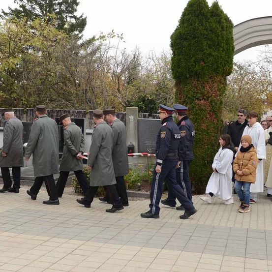 Mehrere Menschen in Uniformen gehen auf einem Friedhof, in der Nähe einer Gruppe von Kindern und Erwachsenen.