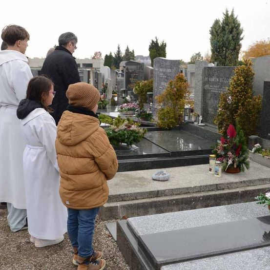 Eine Familie steht an einem Grab in einem Friedhof, mit einem Kind in einer braunen Jacke im Vordergrund und anderen in weißen Gewändern dahinter.
