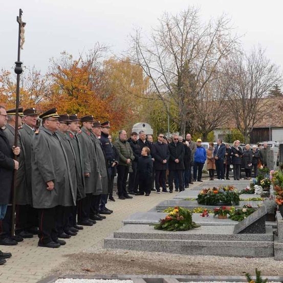 Eine Gruppe von Menschen steht auf einem Friedhof, einige in Uniformen. Im Vordergrund steht ein Kreuz. In der Nähe befinden sich Blumen und Grabsteine. Bäume und ein Gebäude sind im Hintergrund.