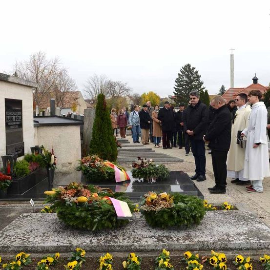Mehrere Menschen sind auf einem Friedhof versammelt, möglicherweise für eine Gedenkfeier. Einige stehen, andere knien. Sie sind in formeller Kleidung gekleidet. Es gibt mehrere Grabsteine und Pflanzen in der Umgebung.