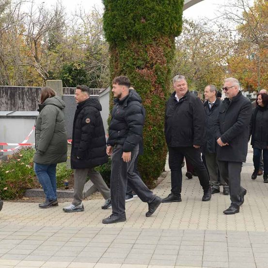 Mehrere Menschen in Jacken gehen in einem Park spazieren und passieren eine hohe Hecke und eine Barriere. Im Hintergrund sind Bäume und Pflanzen zu sehen.