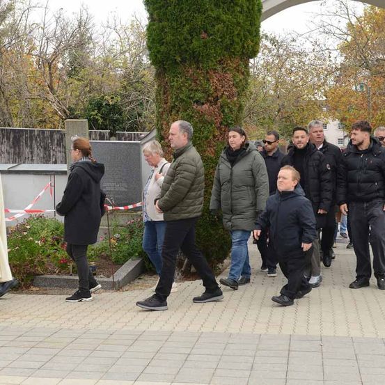 Eine Gruppe von Menschen geht auf einem gepflasterten Weg in der Nähe eines Denkmals, Herbstbäume und ein großer grüner Busch im Hintergrund.