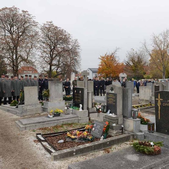 Eine Gruppe uniformierter Personen steht auf einem Friedhof mit verschiedenen Grabsteinen, einige mit Blumen und Kränzen geschmückt. Die Atmosphäre ist feierlich mit Bäumen im Hintergrund.