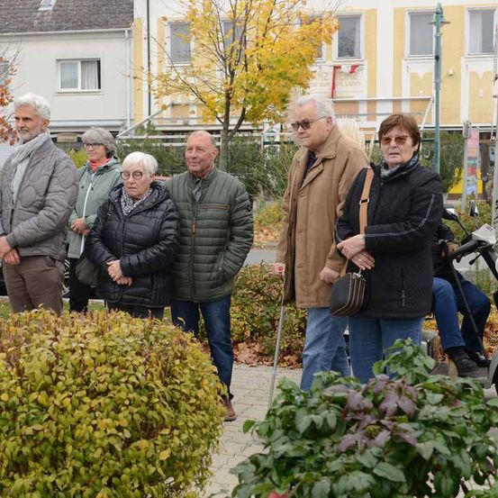 Eine Gruppe älterer Erwachsener steht draußen, möglicherweise bei einer Veranstaltung. Sie sind warm gekleidet, einige tragen Brillen und haben Taschen bei sich. Im Hintergrund befindet sich ein Gebäude mit gelben und weißen Wänden und einem Baum mit gelben Blättern.
