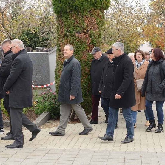 Eine Gruppe von Menschen, die Mäntel tragen, geht in der Nähe einer Gedenktafel, umgeben von Bäumen und Pflanzen in einem Garten.