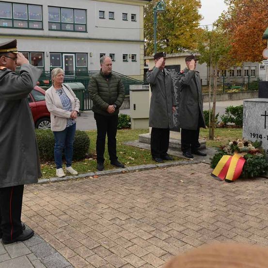 Eine Gruppe von Menschen steht in der Nähe eines Denkmals, darunter zwei uniformierte Männer, die salutieren. Eine Frau und ein Mann stehen hinter ihnen. Das Denkmal hat ein Kreuz und eine Tafel mit dem Jahr 1914.
