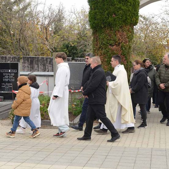 Mehrere Menschen, darunter zwei Priester, gehen gemeinsam auf einem Pfad vor einem Denkmal. Dahinter steht eine schwarze Wand mit einer Tafel, umgeben von Grün.