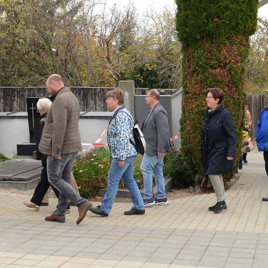 Eine Gruppe von Menschen läuft in einem Park. Eine Frau in der Mitte schaut nach links. Dahinter sind ein Zaun, eine Steinstruktur und ein Baum zu sehen. Der Hintergrund ist unscharf.