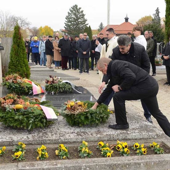 Eine Gruppe von Menschen versammelt sich um ein Grab auf einem Friedhof und legt Kränze und Blumen nieder. Zwei Männer knien sich, um die Kränze anzupassen.