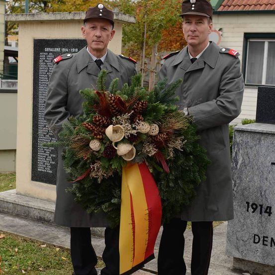 Zwei Männer in Militäruniformen stehen vor einem Denkmal und halten einen Kranz mit Tannenzapfen und einer rot-gelben Schleife. Eine schwarze Tafel listet Namen und Jahre auf, mit 1914 auf einem nahen Stein.