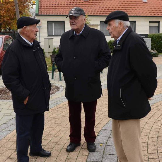 Drei ältere Männer stehen draußen und unterhalten sich. Der Mann auf der linken Seite trägt einen schwarzen Mantel, Hose und Hut. Der Mann in der Mitte trägt einen schwarzen Mantel, rote Hosen und einen schwarzen Hut. Der Mann auf der rechten Seite trägt einen schwarzen Mantel und eine beige Hose. Hinter ihnen steht ein Haus mit rotem Dach und ein geparktes rotes Auto.