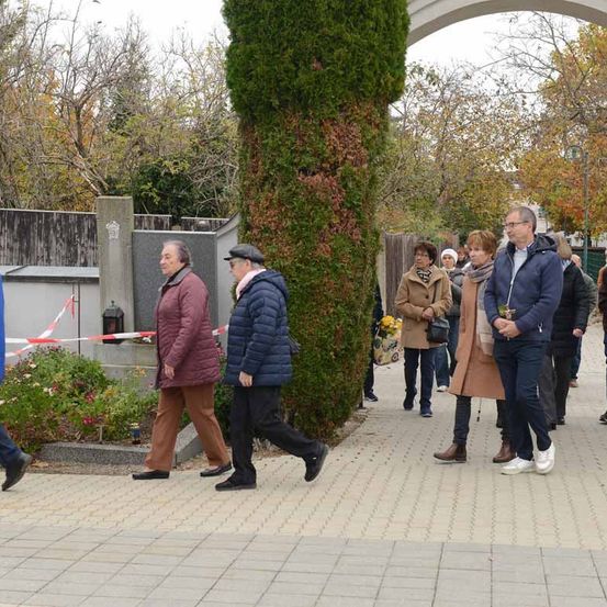 Eine Gruppe von Menschen in Winterkleidung geht an einem Grab vorbei, das mit einem roten Band gekennzeichnet ist, auf einem gepflasterten Weg mit Bäumen und einem Holzzaun im Hintergrund.