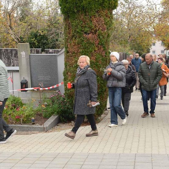 Eine Gruppe von Menschen geht an einem Grab vorbei, das von einem großen Baum und einem Gedenkstein umgeben ist.