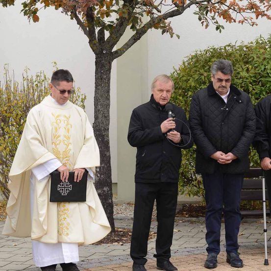 Ein Priester mit einem Buch und drei Männer stehen draußen in der Nähe eines Baumes und einer weißen Wand.