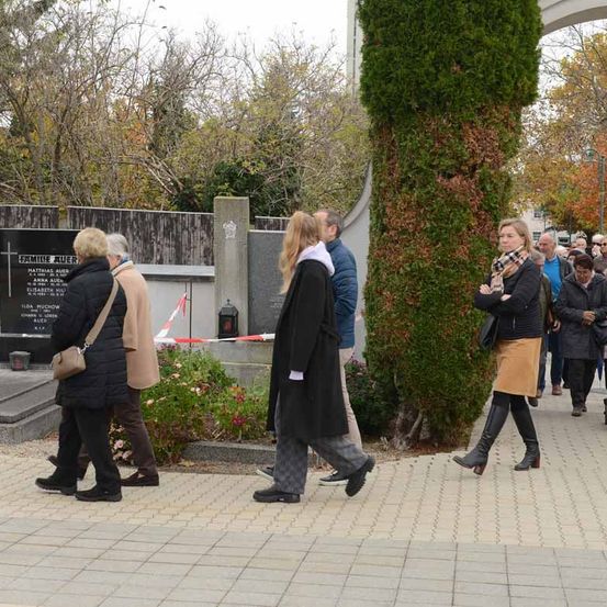Eine Gruppe von Menschen geht auf einem gepflasterten Bereich in der Nähe eines Grabes. Einige tragen Jacken und Stiefel. Eine Frau mit einem Schal schaut auf eine Gedenktafel.