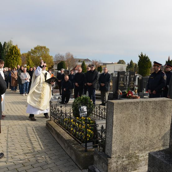 Ein Priester in einer weißen Robe hält ein Buch und eine Glocke auf einem Friedhof mit Blumen und Grabsteinen, wobei eine Menschenmenge hinter ihm steht.