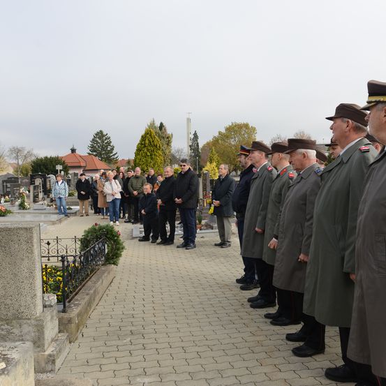 Eine Gruppe uniformierter Personen steht in einer Reihe auf einem Friedhof. Dahinter stehen einige Leute und unterhalten sich in der Nähe der Gräber.