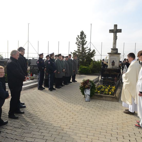 Eine Gruppe von Menschen steht auf einem Friedhof mit einem Kreuz und Blumen vor einem Grabstein. Sie tragen Uniformen und Mützen.