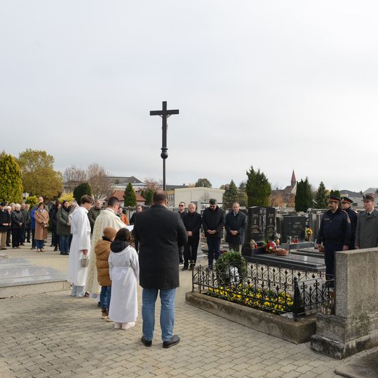 Eine Gruppe von Menschen steht in einem Friedhof mit Blumen und einem Kreuz. Einige sind in Uniform und andere in Mänteln. Einige Menschen beten.