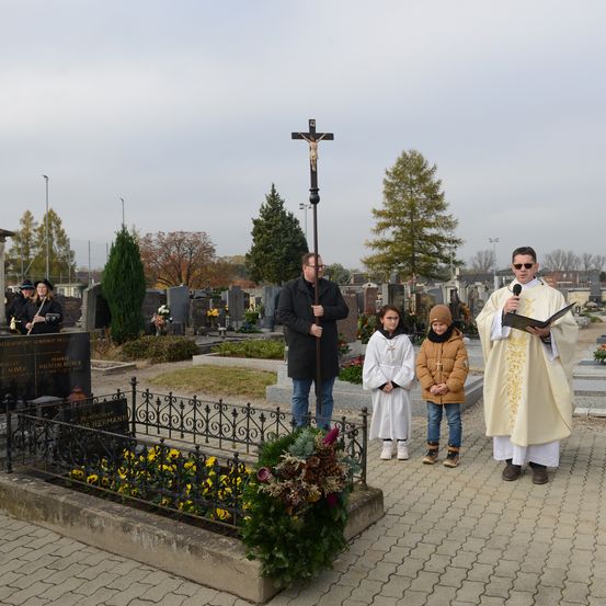 Ein Priester in einer weißen Robe spricht in ein Mikrofon und steht auf einem Friedhof mit einem Kreuz und Blumen. Hinter ihm hält ein Mann in einem schwarzen Mantel einen Gehstock. Zwei Kinder und eine Frau in Schwarz stehen in der Nähe.
