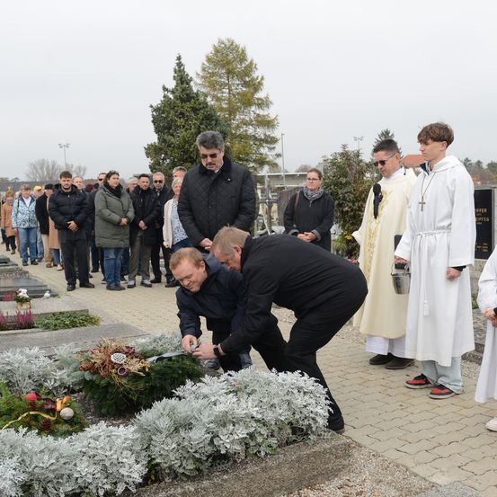 Zwei Männer legen einen Kranz auf ein Grab nieder, während andere zusehen. Priester stehen in der Nähe, einer hält einen kleinen Eimer.
