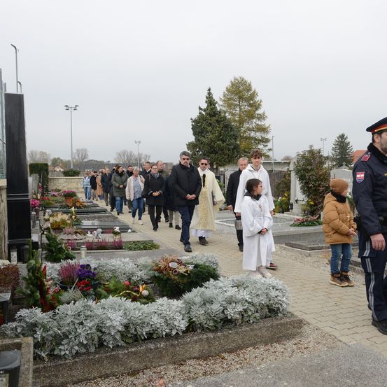 Ein Trauerzug geht durch einen Friedhof, umgeben von Blumen und Bäumen. Zwei Priester in weißen Gewändern führen die Gruppe an. Ein Kind steht zur Seite, trägt einen braunen Mantel und einen Hut. Ein Polizist ist ebenfalls anwesend.
