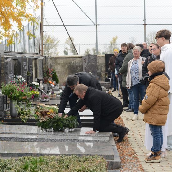 Mehrere Menschen versammeln sich auf einem Friedhof und legen Blumen auf Grabsteine. Zwei Männer knien, einer mit einem Kranz. Zuschauer stehen dahinter und ein junges Kind beobachtet.