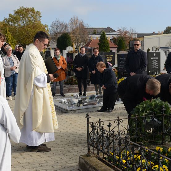 Ein Priester leitet eine Zeremonie auf einem Friedhof, ein Junge legt einen Kranz nieder. Menschen stehen um ein Grabmal.
