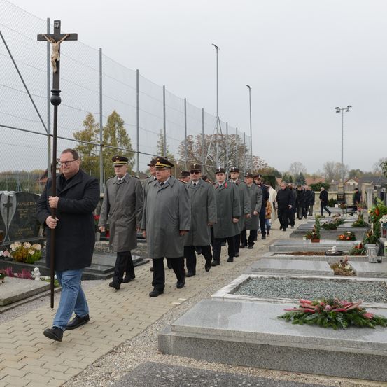 Eine feierliche Prozession von Personen, gekleidet in lange graue Mäntel und Hüte, marschiert an einem Kreuz auf einem gepflasterten Weg vorbei in Richtung eines Friedhofs mit Grabsteinen, die mit Blumen und Kreuzen geschmückt sind.
