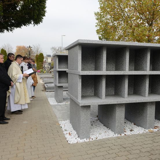 Ein Priester liest eine Trauerrede bei einer Beerdigung, steht neben einer großen, grauen, modularen Bestattungsstruktur mit mehreren Fächern, in einem Friedhof mit Bäumen und gepflasterten Wegen.