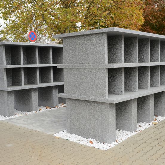 Ein großes graues Betongebäude mit mehreren Nischen, das einem Mausoleum ähnelt, steht auf einem Ziegelweg. Der Bereich ist von Bäumen mit gelben und orangefarbenen Blättern umgeben. Ein rotes und weißes Schild befindet sich auf dem Gebäude.