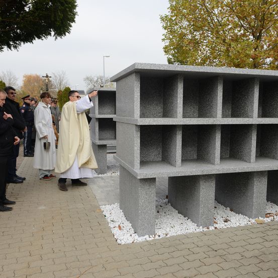 Ein Priester führt eine Zeremonie vor einer großen, grauen, modularen Struktur durch. Er hält eine kleine Glocke in der Hand, und mehrere Personen stehen in der Nähe, gekleidet in formelle Kleidung. Bäume mit gelben Blättern und ein bewölkter Himmel bilden den Hintergrund.