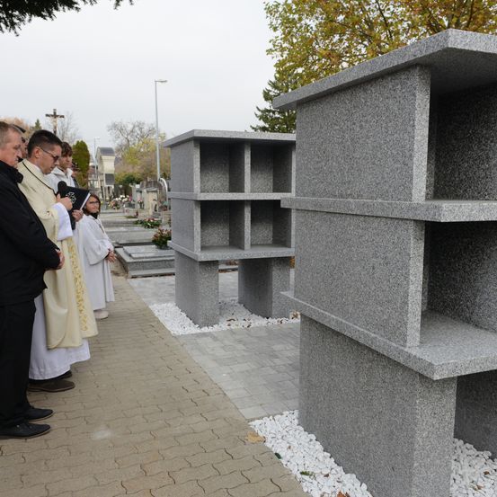Eine Gruppe von Menschen in formeller Kleidung steht an einem Friedhof, möglicherweise bei einem Beerdigungsgottesdienst. Das Grab ist aus Granit mit mehreren Fächern.