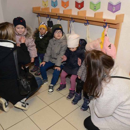 Eine Gruppe von Kindern sitzt auf einer Bank in einem Raum. Sie tragen Winterkleidung. Zwei Frauen sitzen vor ihnen auf dem Boden.