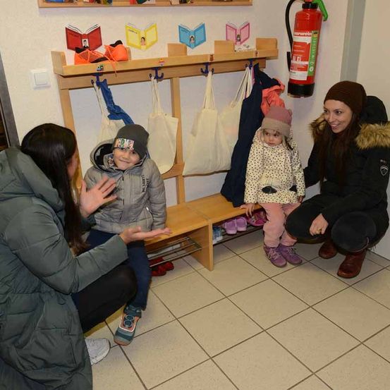 Drei Frauen und zwei Kinder sitzen auf einer Bank in einem Raum mit gefliesten Böden. Ein Feuerlöscher ist an der Wand montiert.