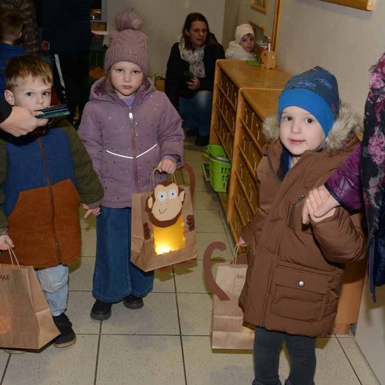 Drei Kinder halten Papiertüten mit einer leuchtenden Affenillustration in einem Flur. Eine Frau sitzt im Hintergrund auf dem Boden.