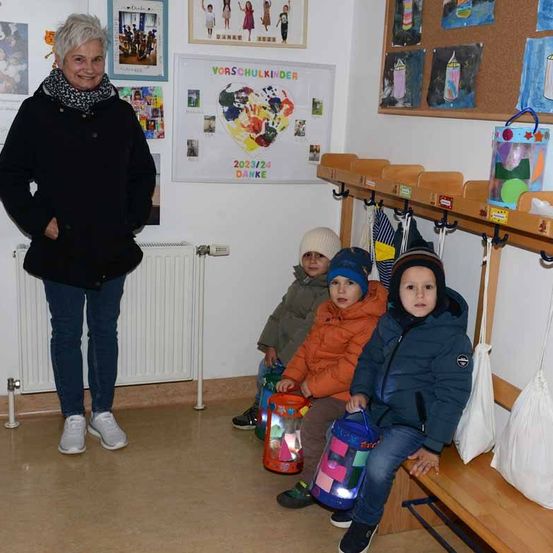 Eine Frau steht mit den Händen auf den Hüften in einem Raum. Drei Kinder sitzen auf einer Bank, jedes hält eine Laterne.