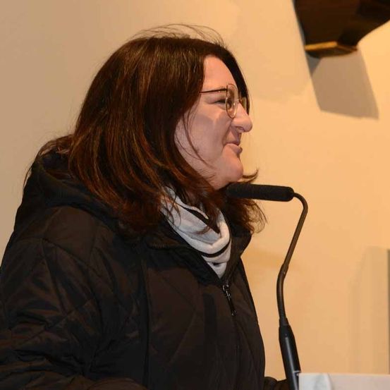 Eine Frau mit braunen Haaren, die eine Brille und eine schwarze Jacke trägt, spricht in ein Mikrofon am Rednerpult.