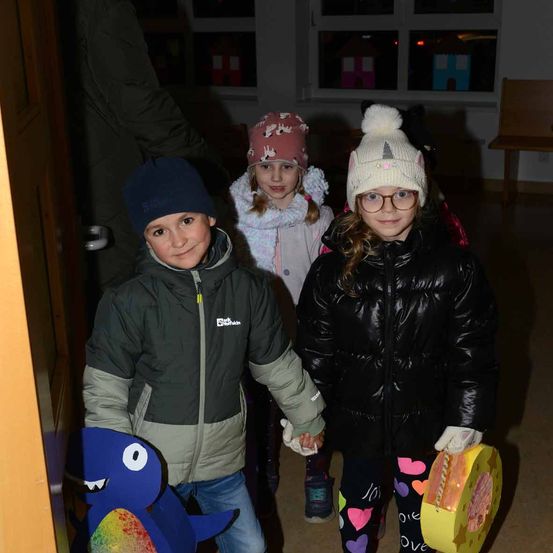 Drei Kinder in Winterkleidung stehen lächelnd nebeneinander. Der Junge auf der linken Seite hält einen blauen Dinosaurier. Hinter ihnen ist ein Raum mit Stühlen und Fenstern zu sehen.