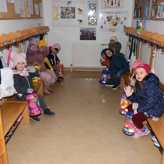 Eine Gruppe von Kindern sitzt auf Bänken in einem Raum mit weißen Wänden, einem Heizkörper und Kunstwerken. Sie tragen Winterkleidung und halten Taschen und lächeln für ein Foto.