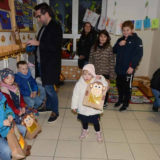 Eine Gruppe von Kindern hält Taschen und steht oder sitzt in einem Raum. Ein Mann in einem schwarzen Mantel steht und hält ein Objekt. Hinter den Kindern steht eine Frau in der Nähe eines Fensters mit einem bunten Banner.